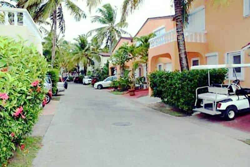 Antigua chiama Italia, terra magica tra cielo e mare 3 Colorato quartiere residenziale con palme, case color pastello e auto sportive lungo una strada tranquilla, perfetto per vacanze rilassanti e soggiorni al mare.