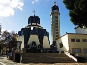 Chiesa moderna con architettura audace e torre campanaria, situata in un contesto urbano con alberi e scalinate, rappresenta un esempio di architettura religiosa innovativa in Italia.