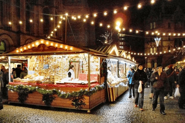 Suggestive Christmas market stall illuminato con luci e decorazioni festive, atmosfera natalizia vivace con visitatori che passeggiano tra le bancarelle in un contesto urbano serale.