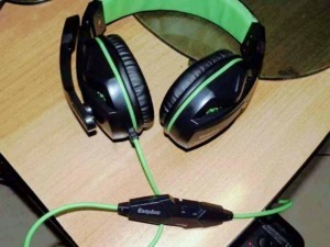 Auricolari gaming con microfono e cavo verde, headset over-ear nere e verdi per gaming e streaming, collocati su un tavolo di legno nero, accessori elettronici gaming.