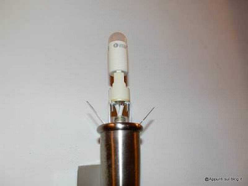 Lampada led impressa in un tubo di metallo, accesa, vicino alla parete bianca.