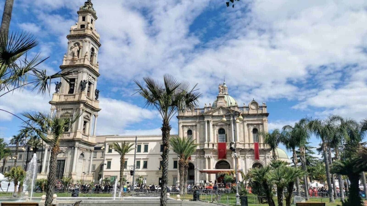 Immagine della Basilica di San Paolo, chiesa storica e simbolo di Napoli, circondata da palme e giardini, con un cielo azzurro e nuvole bianche sullo sfondo.