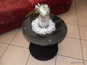 Vaso con fiori freschi e dotto in vetro su un tavolino tondo in rattan nero con coperta di pizzo, arredamento moderno e elegante per soggiorno.