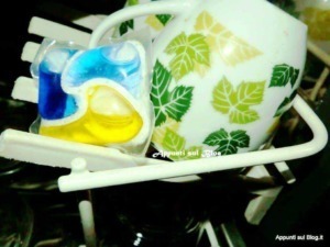 Vaschetta con piatti e tazze in ceramica decorata con foglie verdi e gialle, con saponi colorati, posizionata su un supporto bianco, ideale per l'uso quotidiano in cucina o in casa.