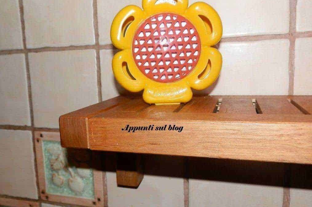 Fiore in plastica giallo e rosso, decorativo e colorato, posizionato sopra mensola di legno, su parete di piastrelle beige, con scritto "Appunti sul blog" sul legno.