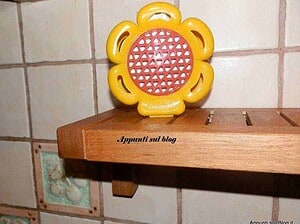 Fiore in plastica giallo e rosso, decorativo e colorato, posizionato sopra mensola di legno, su parete di piastrelle beige, con scritto "Appunti sul blog" sul legno.