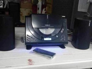 Alt text:Sistema stereo con lettore CD e due altoparlanti neri posizionati su un tavolo bianco, con telecomando davanti.
