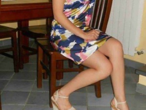 Relaxed woman sitting on a wooden chair in a colorful summer dress and high heels, indoor setting, casual and stylish vibe.
