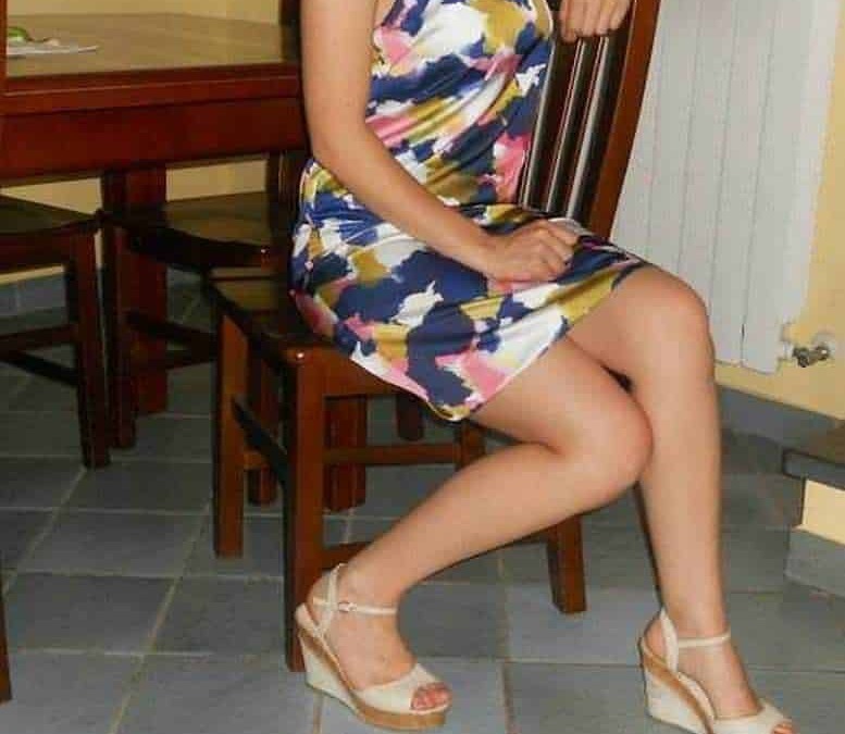Relaxed woman sitting on a wooden chair in a colorful summer dress and high heels, indoor setting, casual and stylish vibe.