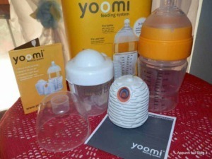 Pompa alimentazione biberon Yoomi, sistema di riscaldamento automatico per latte, accessori per allattamento, pratici e innovativi prodotti per neonati, sul tavolo con caraffa e scatola.
