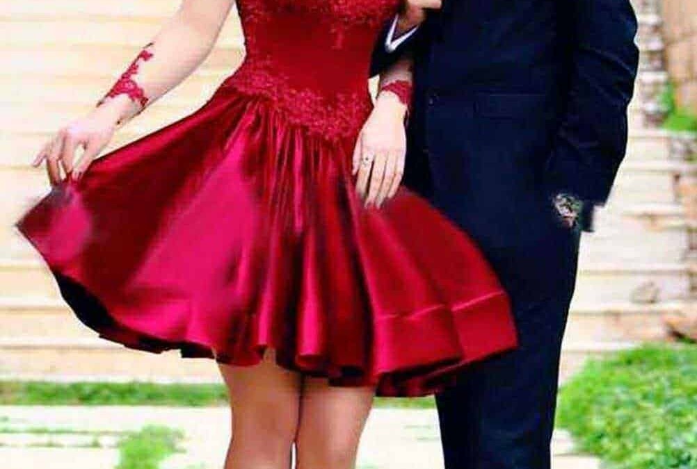 Elegante coppia innamorata con donna in abito rosso da cerimonia e uomo in completo nero, passeggiata romantica su marciapiede all'aperto, sfondo urbano.