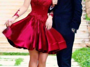 Elegante coppia innamorata con donna in abito rosso da cerimonia e uomo in completo nero, passeggiata romantica su marciapiede all'aperto, sfondo urbano.