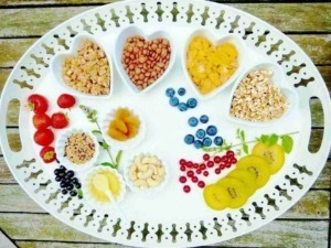 Fragole, mirtilli, kiwi, e frutta secca in ciotole di cuore, con altri frutti e bacche disposti su un vassoio bianco per una colazione sana e colorata.