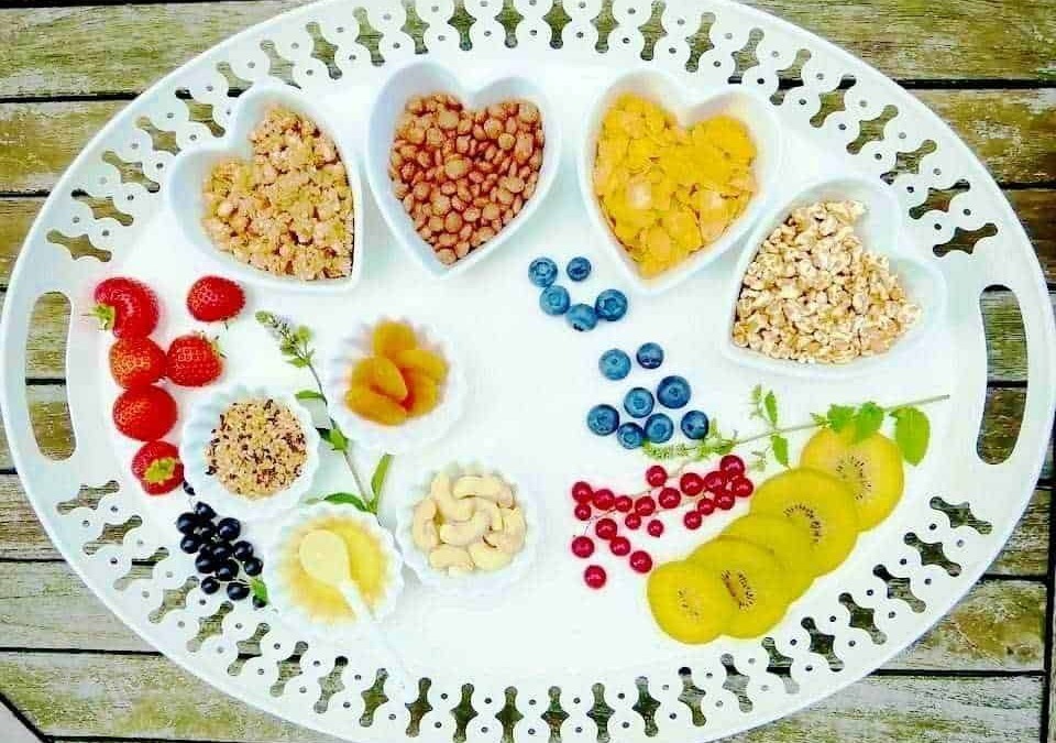 Fragole, mirtilli, kiwi, e frutta secca in ciotole di cuore, con altri frutti e bacche disposti su un vassoio bianco per una colazione sana e colorata.