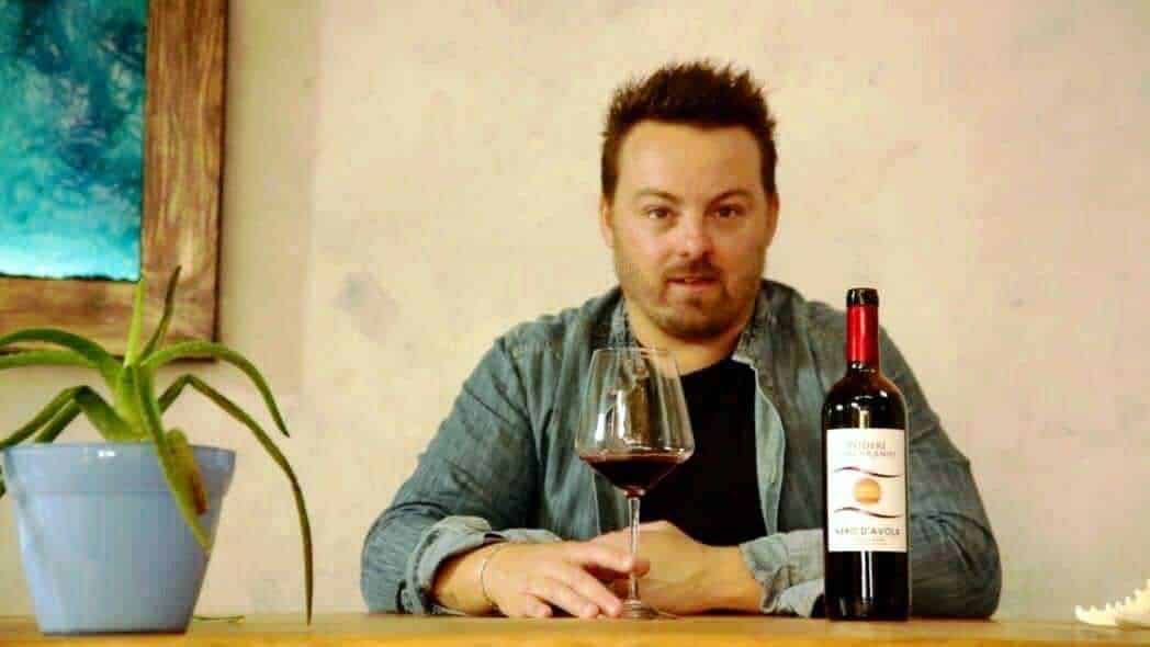 Un uomo che beve vino rosso in un ambiente domestico con una bottiglia di vino, un bicchiere e una pianta sul tavolo, atmosfera accogliente e rilassante.