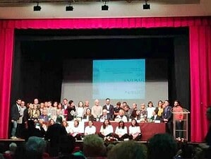 Riconoscimento di un gruppo di persone sul palco di un teatro durante un evento ufficiale, con schermo e pubblico presente.