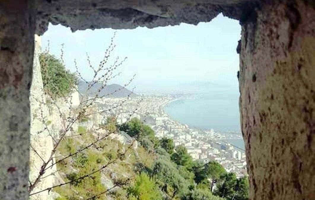 Veduta panoramica della costa e della città vista attraverso una fessura in una torre di pietra, con vegetazione verde e mare sullo sfondo, in un ambiente storico e naturale di grande fascino.