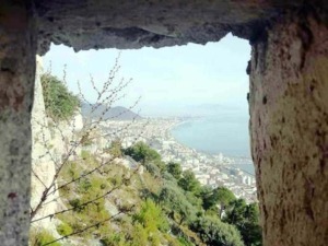 Veduta panoramica della costa e della città vista attraverso una fessura in una torre di pietra, con vegetazione verde e mare sullo sfondo, in un ambiente storico e naturale di grande fascino.