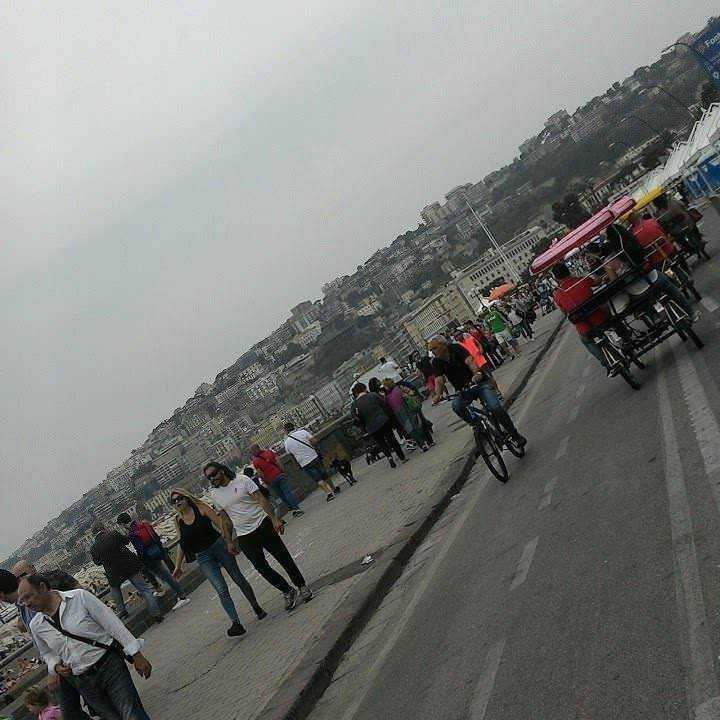Pedalare in bicicletta: perché fa bene. Passeggiata sul lungomare con pedoni, ciclisti e carri di cibo sotto un cielo nuvoloso, con la città sullo sfondo