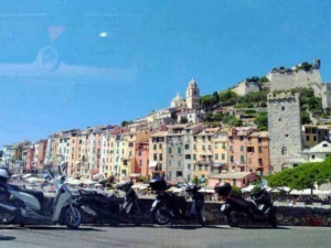Colorato panorama di una pittoresca cittadina ligure con case fantasiose e un castello antico sullo sfondo, vista dalla strada con scooter parcheggiati in primo piano.