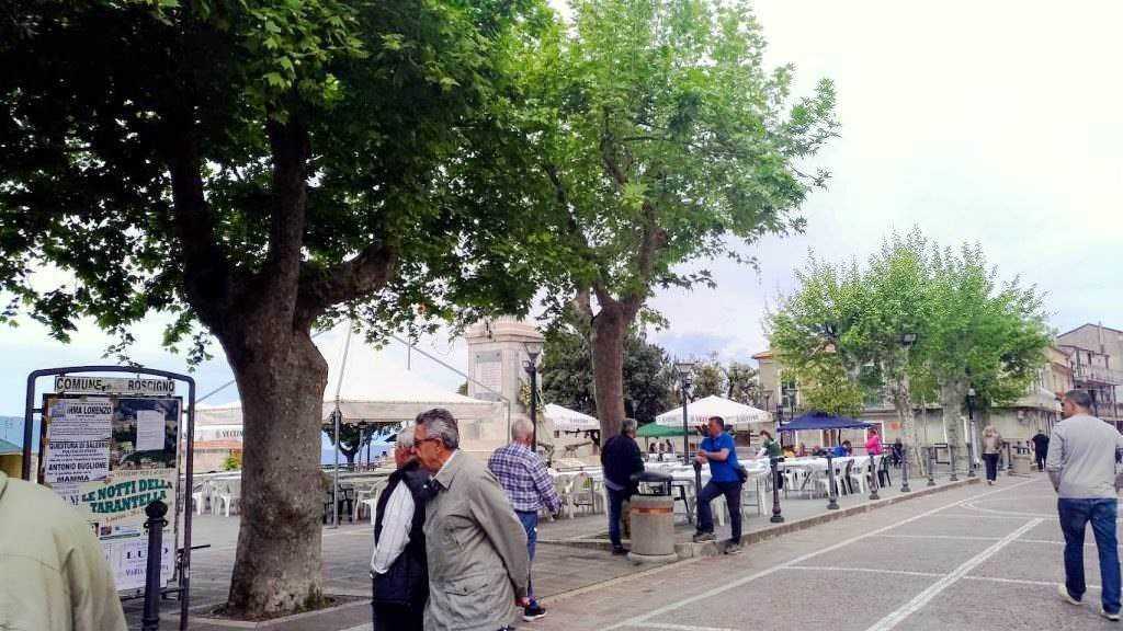 Passeggiata nel centro di Rossano, con albero al centro e persone che si godono la domenica in un'atmosfera tranquilla e vivace, tra negozi e panchine.