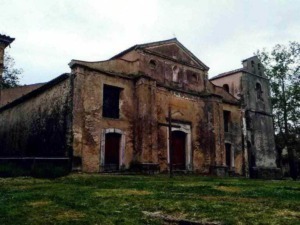 Vecchia chiesa in rovina di epoca storica, con muri in mattoni rossi e dettagli architettonici decadenti, circondata da alberi verdi e un cortile erboso sotto un cielo nuvoloso.