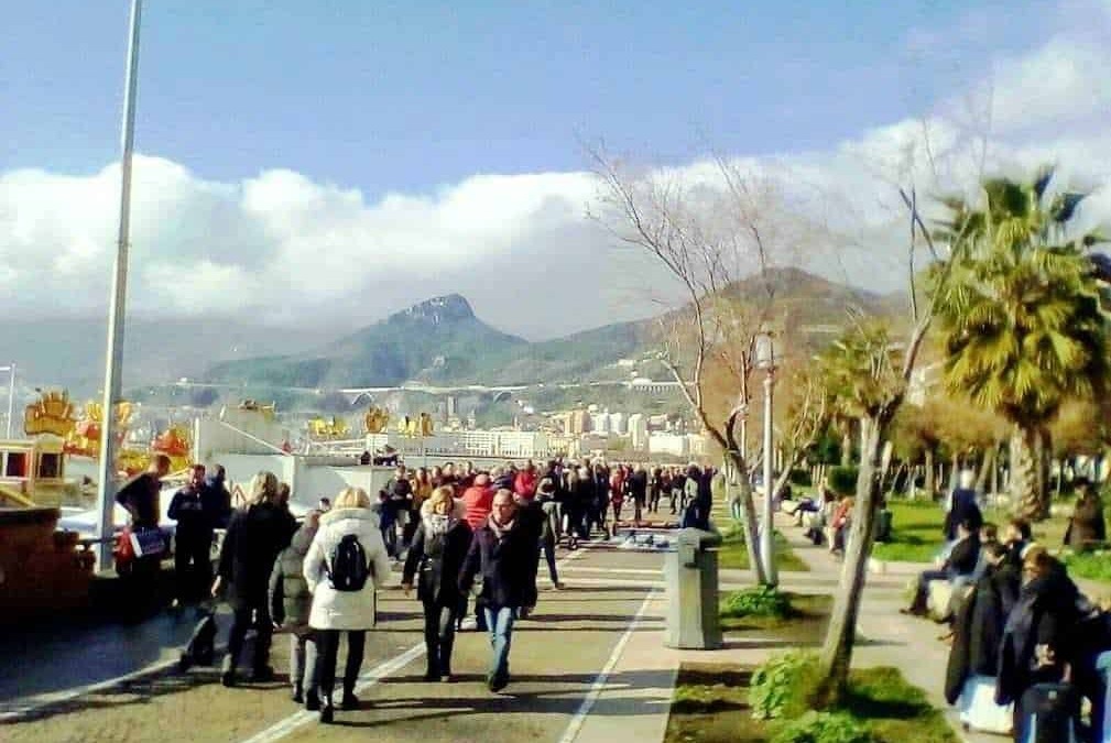 Passeggiata sul lungomare di Napoli con vista sul Vesuvio, persone che si godono una giornata di sole, ambiente vivace e accogliente nel centro città.
