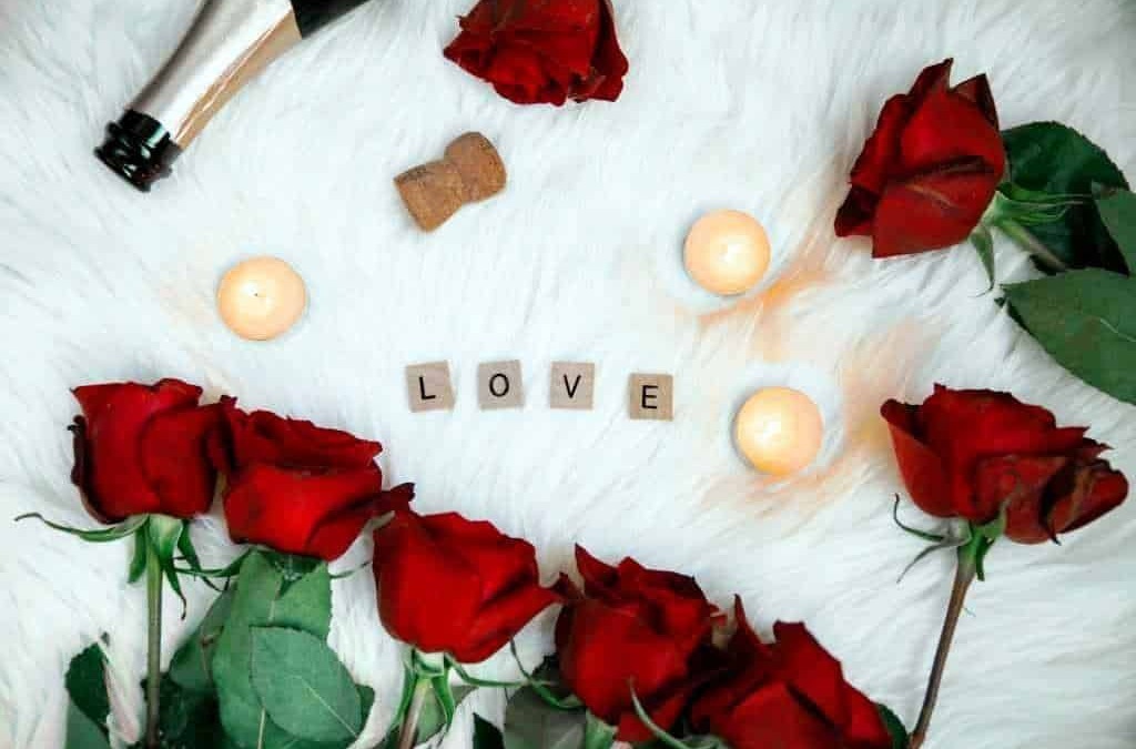 Roses rosse, candele accese, e lettere a formare la parola "LOVE" su un morbido sfondo bianco, creando un'atmosfera romantica e suggestiva per occasioni speciali o momenti di affetto.
