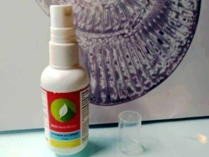 Spray di vitamina D3 NutrientWise in bottiglia bianca con etichetta rossa, posizionata su una superficie trasparente, con un bicchiere di plastica trasparente e sfondo astratto colorato.