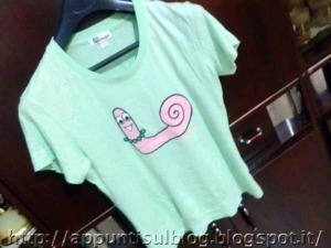 Una maglietta verde con personaggio cartoon a forma di lumaca sorridente, ideale per fan di personaggi divertenti e accessori di moda colorati.