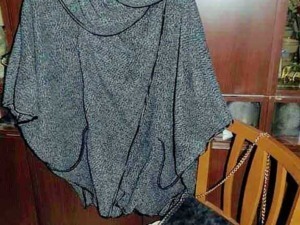 Oversized grey knit poncho with black piping, hanging on a wooden hanger.
