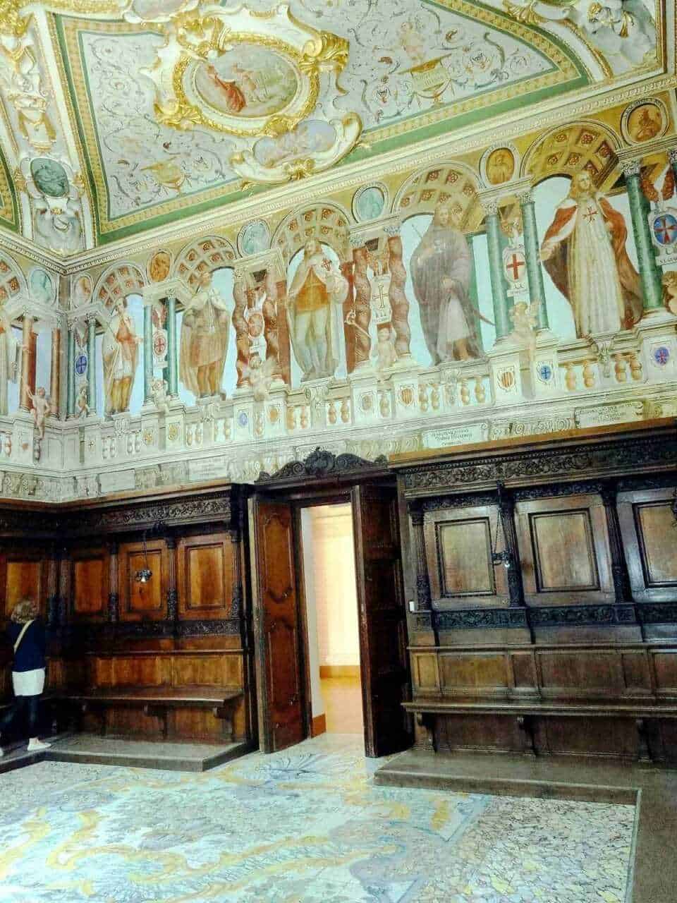 Faro di giustizia con dipinti sacri sul soffitto, pareti adornate con affreschi religiosi e arredi in legno antico in stile barocco, ambiente interno di una chiesa o cappella storica italiana.