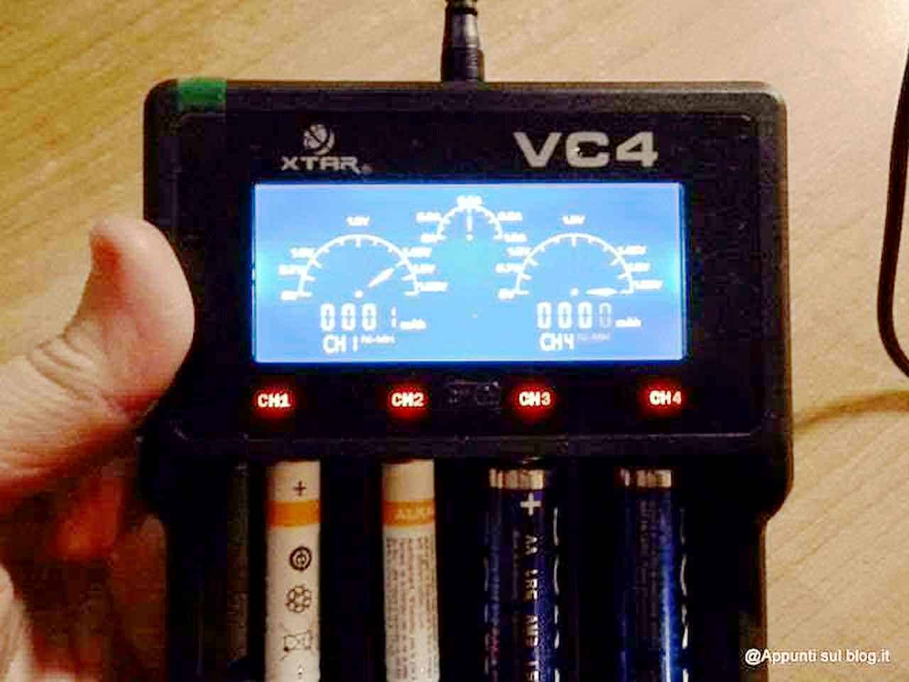 Caricabatteria pile con funzione zero volt: Relay screenshot of a multichannel digital voltmeter with a blue backlit display showing four analog signals. The device is held in a person's hand, with batteries visible at the bottom. The device is labeled "VC4" and has four channels, used for measuring electrical voltage.