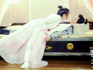 Ragazza vestita con abiti tradizionali asiatici che si avvicina a un uomo sdraiato a letto, scena in stile storico o cinematografico con dettagli eleganti e ambientazione orientale.