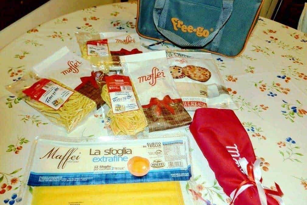 Pacchetti di pasta Maffei e accessori per la cucina su tavolo con tovaglia floreale, sacchetto rosso e borsa da viaggio.