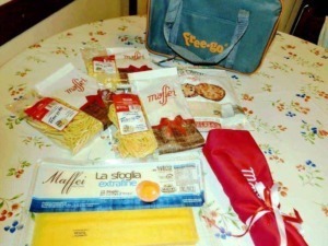 Pacchetti di pasta Maffei e accessori per la cucina su tavolo con tovaglia floreale, sacchetto rosso e borsa da viaggio.