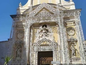 Majestic church facade with intricate baroque architecture and artistic sculptures, under bright blue sky, featuring decorative scaffolding for restoration work, located on Striano street in Italy.
