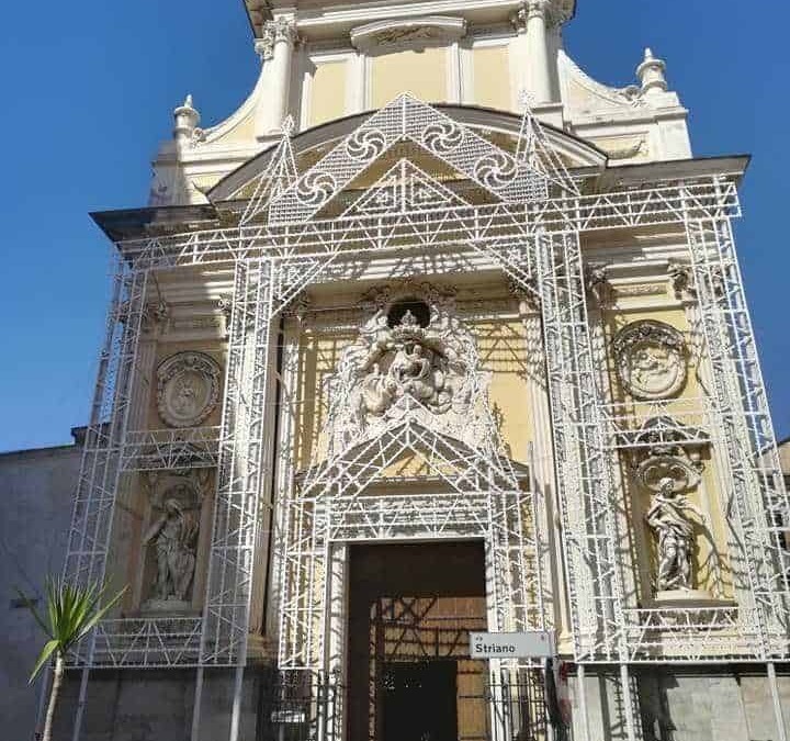 Majestic church facade with intricate baroque architecture and artistic sculptures, under bright blue sky, featuring decorative scaffolding for restoration work, located on Striano street in Italy.