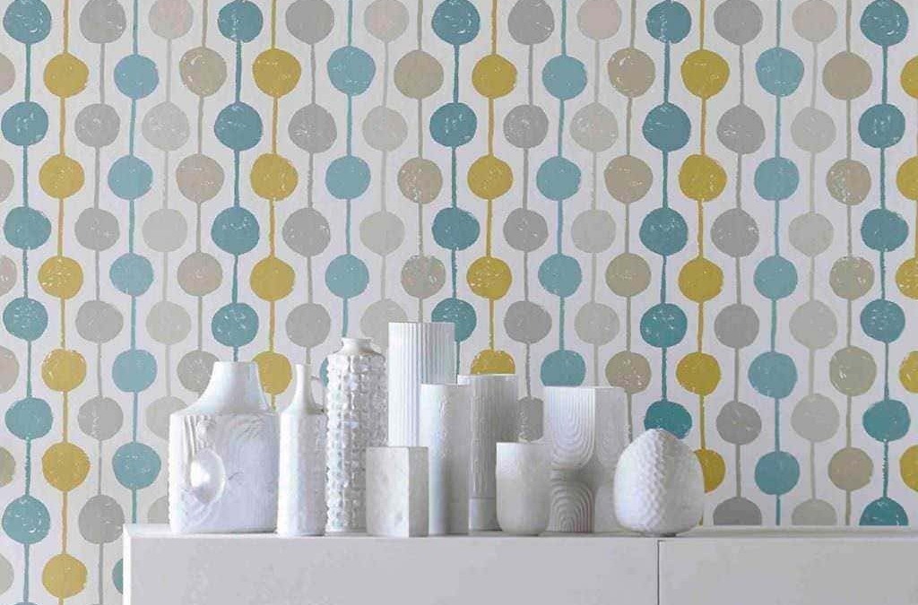 Colorata carta da parati con motivi a cerchi in toni pastello blu, grigio e giallo, decorando un muro interno moderno. Vasi bianchi vintage sono disposti su una superficie bianca minimalista.
