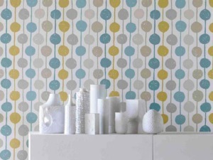 Colorata carta da parati con motivi a cerchi in toni pastello blu, grigio e giallo, decorando un muro interno moderno. Vasi bianchi vintage sono disposti su una superficie bianca minimalista.