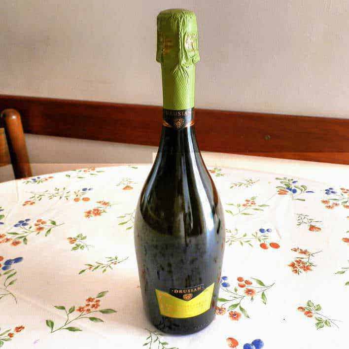 Bottiglia di spumante con effervescenza e capsula verde su tovaglia floreale, ideale per festeggiamenti e occasioni speciali.