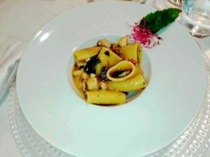 Farfalle con seppie e salsa al nero di seppia decorata con erbe fresche e fiori commestibili, presentata su piatto elegante in ristorante di alta cucina.