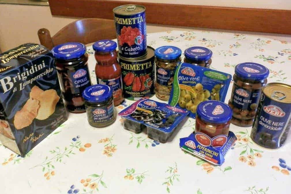 Pomodori pelati in scatola, conserve di olive nere denocciolate, sottaceti di cipolle e peperoncini, e snack di olives greche e sott'olio su tavolo con tovaglia floreale.