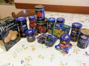Pomodori pelati in scatola, conserve di olive nere denocciolate, sottaceti di cipolle e peperoncini, e snack di olives greche e sott'olio su tavolo con tovaglia floreale.