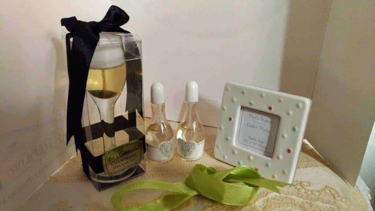 Cofanetto regalo con bottiglia di vino decorato con fiocco nero, due profumi in bottiglia, cornice portafoto con decorazioni colorate, dettagli regalo su tovaglia beige.