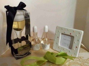 Cofanetto regalo con bottiglia di vino decorato con fiocco nero, due profumi in bottiglia, cornice portafoto con decorazioni colorate, dettagli regalo su tovaglia beige.