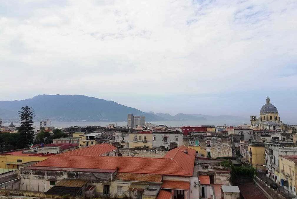 Vista panoramica di Napoli con il Vesuvio sullo sfondo e il Golfo di Napoli, mostra edifici storici e il campanile di una chiesa, cielo nuvoloso, atmosfera urbana e naturale in equilibrio.