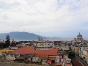 Vista panoramica di Napoli con il Vesuvio sullo sfondo e il Golfo di Napoli, mostra edifici storici e il campanile di una chiesa, cielo nuvoloso, atmosfera urbana e naturale in equilibrio.