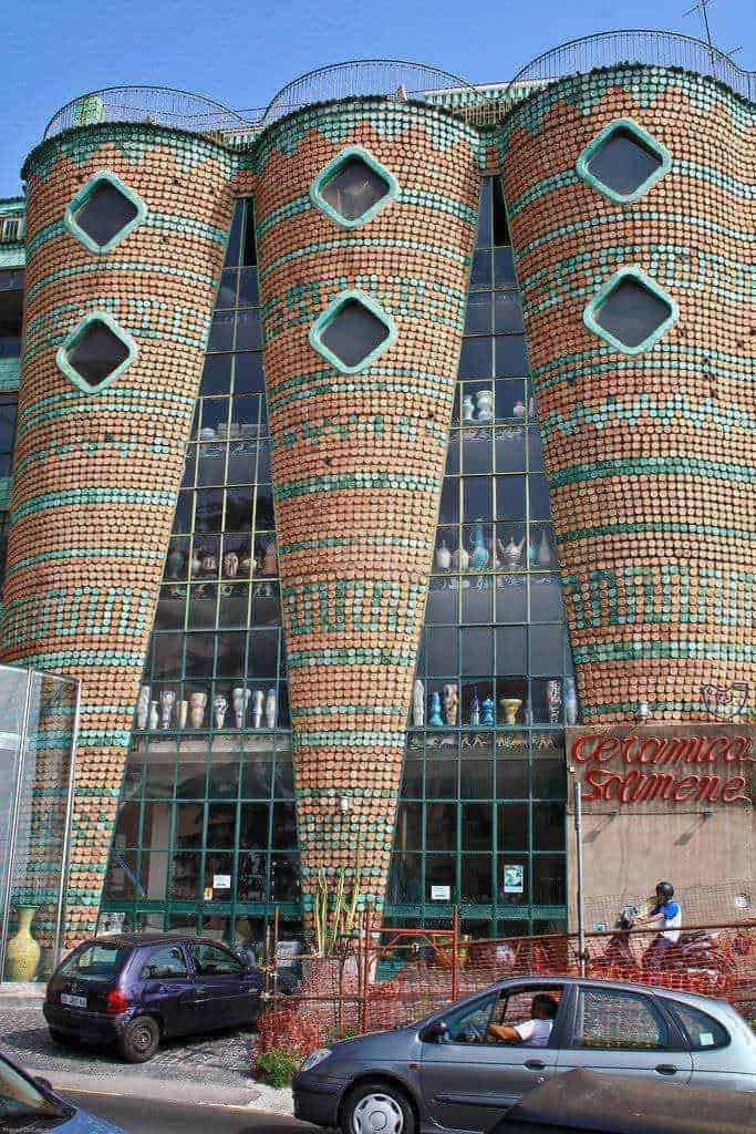 Una facciata di un edificio moderno decorata con mosaici di ceramiche colorate, con grandi vasi di terracotta e vetrate che riflettono il cielo. La struttura si chiama "Ceramica Ramena" ed è un esempio di architettura innovativa e arte urbana.