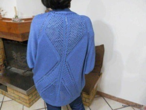 Lei, indossando un maglione blu con dettagli traforati, si trova in un ambiente domestico con un camino in mattoni sullo sfondo.